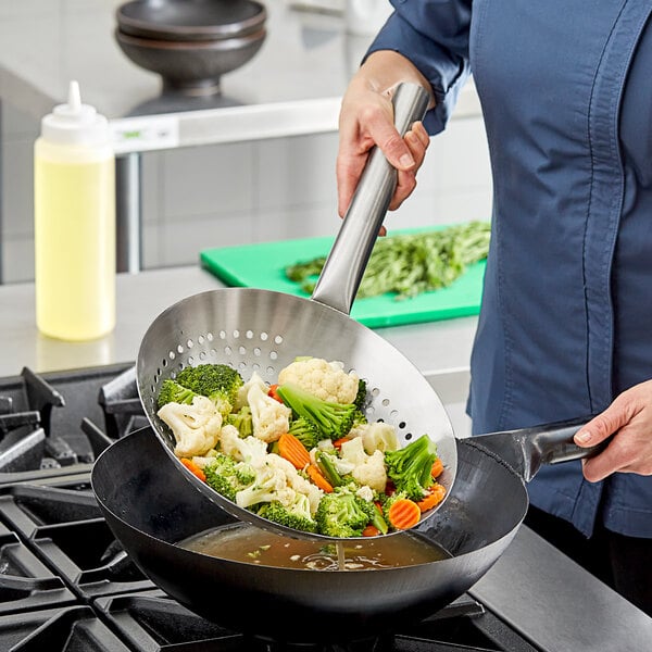 A person using an Emperor's Select stainless steel wok skimmer to cook vegetables in a wok.