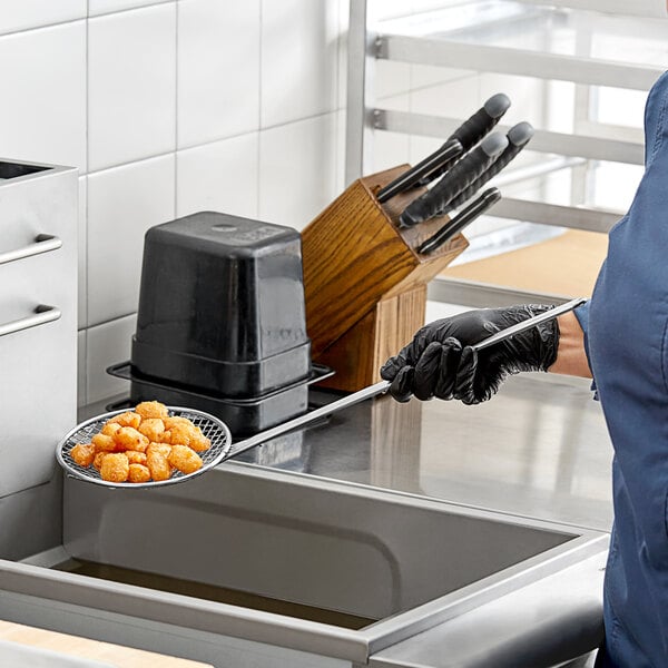 A woman in a blue uniform using a Choice reinforced metal mesh skimmer to hold food over a pan.