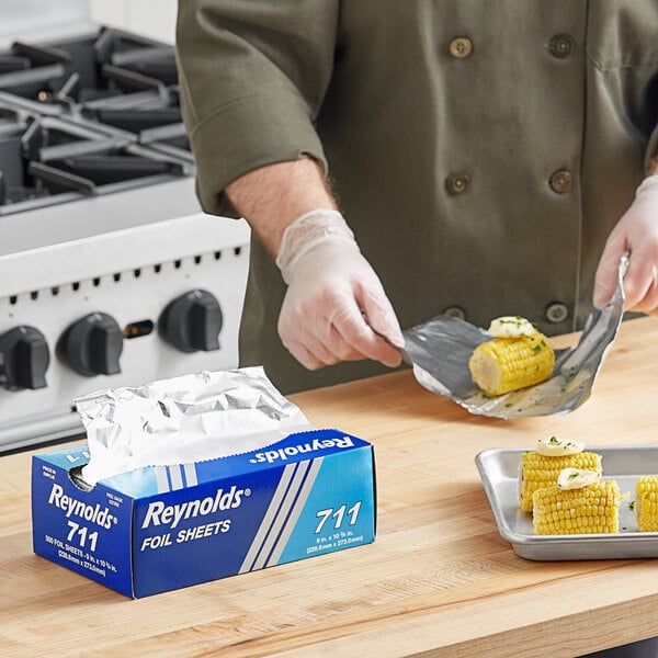 A person wearing gloves and using a Reynolds foil sheet to cut corn on the cob.