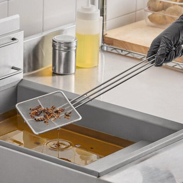 A square fine mesh tinned French fry skimmer being used to remove debris from a fryer.
