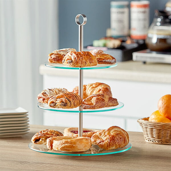 An Acopa three tiered glass and stainless steel display stand with pastries on a table.