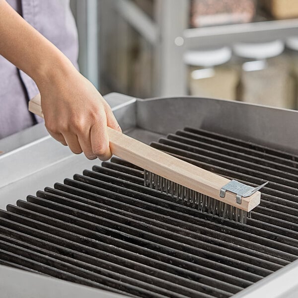 A person's hand holding an American Metalcraft wooden wire brush over a grill.