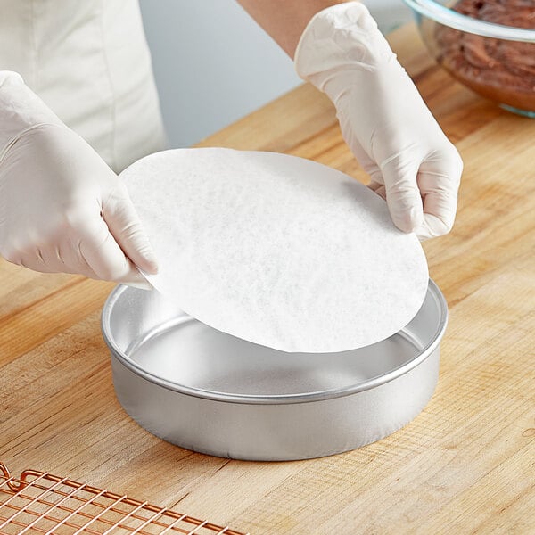 A person wearing gloves places a round white Baker's Lane parchment paper in a round metal pan.