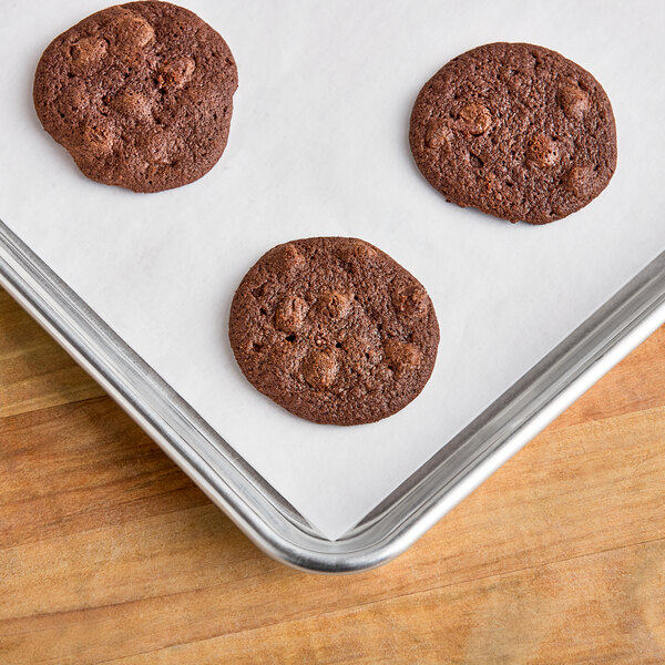 A full size silicone coated parchment paper sheet lining a baking tray with three chocolate cookies on top.