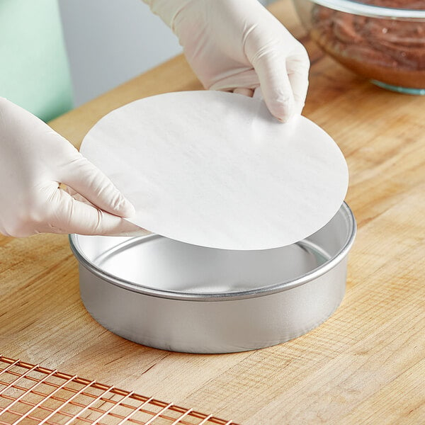 A gloved hand placing a Baker's Lane parchment paper liner in a round metal pan.