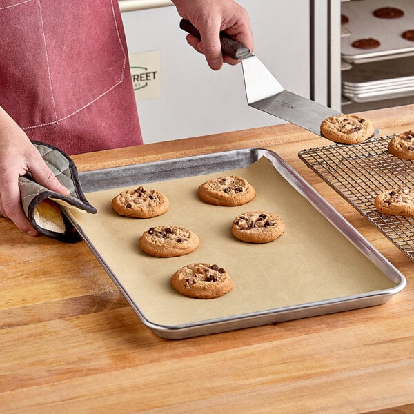 A hand using a spatula to put a cookie on a tray lined with Baker's Lane parchment paper.