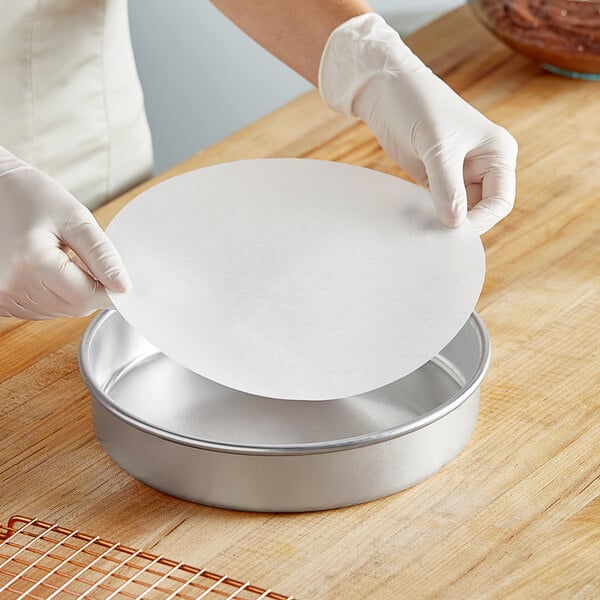 A person wearing gloves placing a Baker's Lane dry wax parchment liner in a round metal pan.