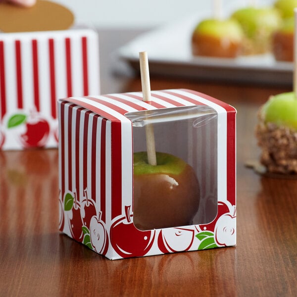A red and white striped Baker's Lane candy apple box with a window and a green candy apple inside.