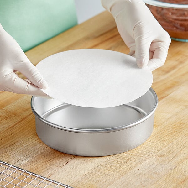 A gloved hand placing a round white Baker's Lane pan liner over a round metal pan.