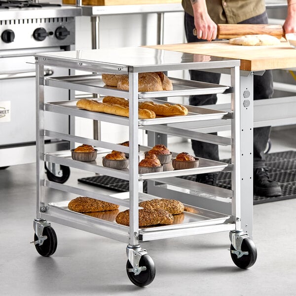 A metal undercounter bun and sheet pan rack with a stainless steel top and side channel, holding trays of baked goods.