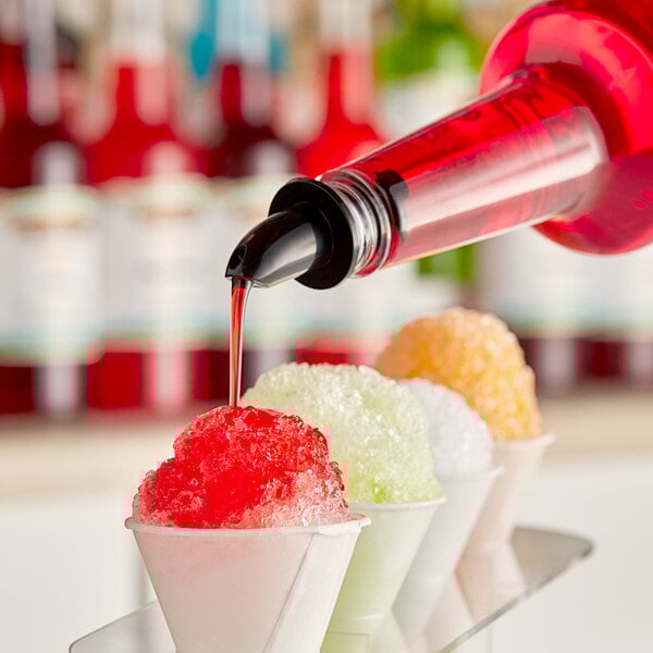 A bottle of red syrup being poured onto a cup of shaved ice, with other colorful shaved ice cups in the background.