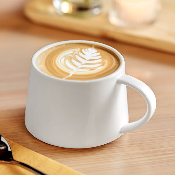 An Acopa Lore bright white porcelain cup filled with coffee with a leaf design in the foam.