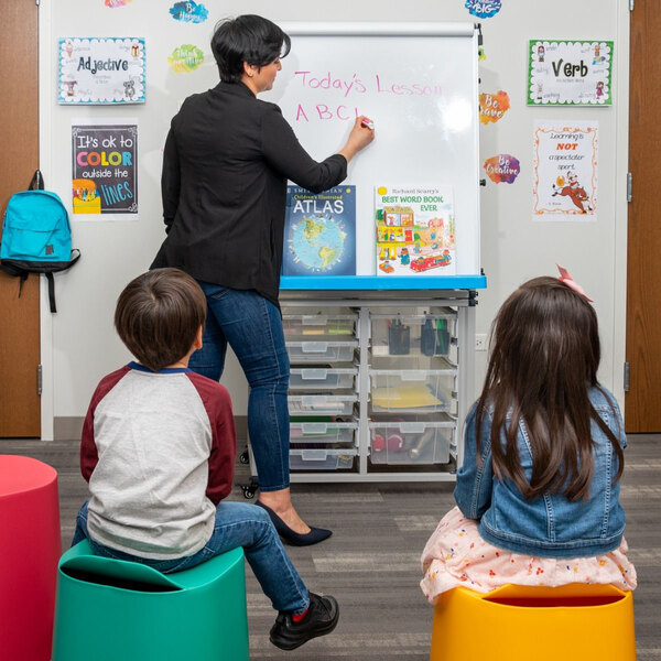 A woman teaching children in a classroom using a Luxor white steel mobile modular teacher dry erase easel.