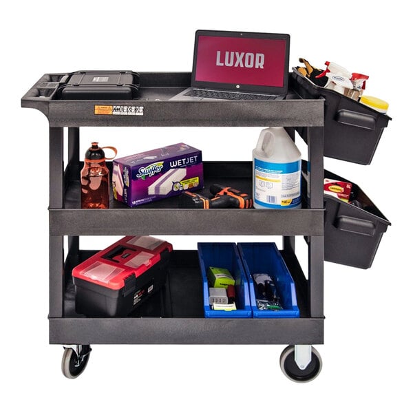 A black Luxor plastic utility cart with two black outrigger bins and cleaning supplies on the shelves.