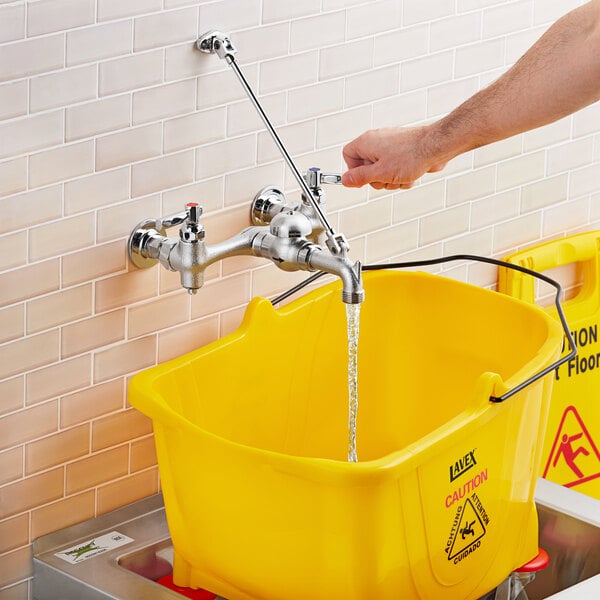 A person using an American Standard wall-mount utility faucet to wash a yellow bucket with a yellow handle.