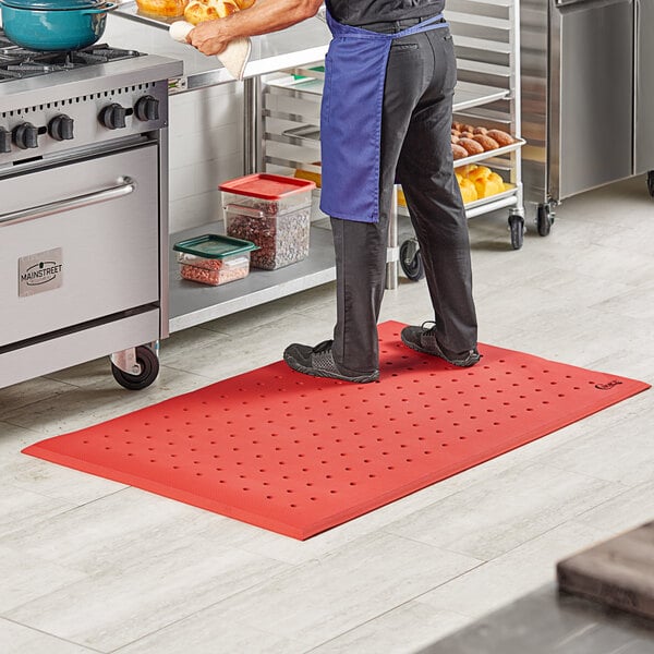 A person in a blue apron standing on a red Choice anti-fatigue floor mat in a professional kitchen.