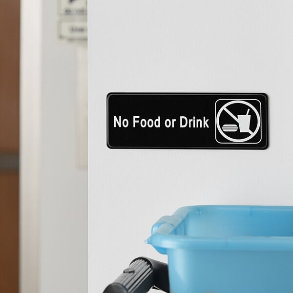 A Lavex black and white sign on a wall above a blue plastic container that says "No Food Or Drink"