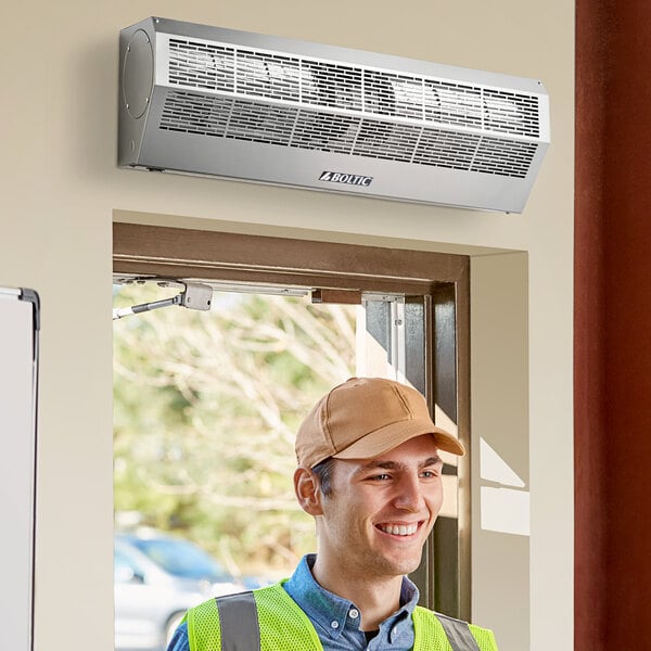 A man in a yellow vest and hard hat standing next to a Boltic stainless steel air curtain over a door.