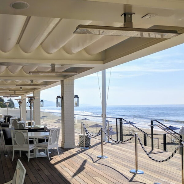 A white electric outdoor patio heater mounted on the ceiling of a covered deck overlooking a beach.