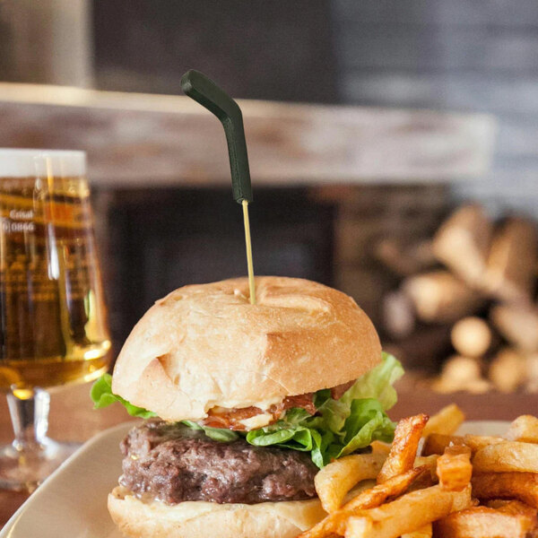 A hamburger with a bamboo cocktail pick shaped like a hockey stick inserted into the top bun, served with fries and a glass of beer in the background.