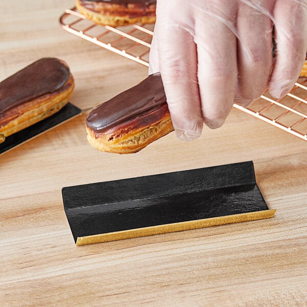 A person holding a chocolate covered eclair on a black and gold rectangular dessert board.