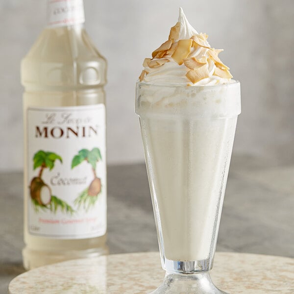 A milkshake with whipped cream and coconut flakes in front of a bottle of Monin Premium Coconut Flavoring syrup.