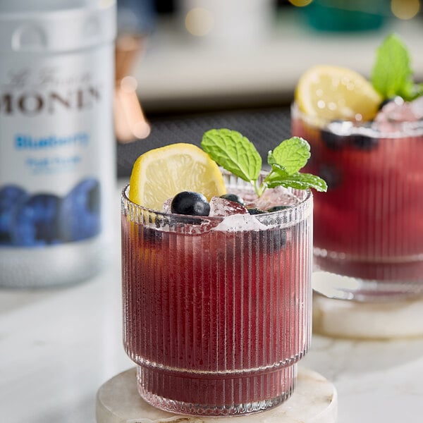 Two glasses of Monin Blueberry Lemonade garnished with mint and blueberries on a marble table with a Monin Blueberry Fruit Puree bottle.