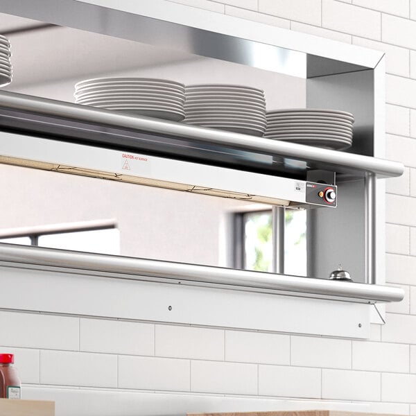 A ServIt strip warmer mounted on a stainless steel shelf above plates in a professional kitchen.