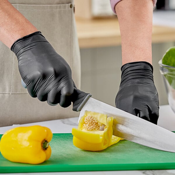 A person wearing black Noble Products disposable gloves cuts a yellow bell pepper on a green surface.