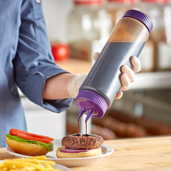 A hand using a Vollrath Traex clear squeeze bottle with purple cap to pour sauce on a burger.