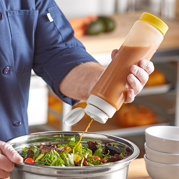 A hand using a Vollrath Traex clear squeeze bottle with white and yellow caps to pour brown sauce onto a bowl of salad.