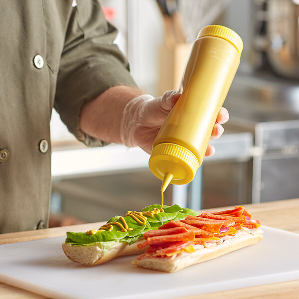 A hand using a Vollrath Traex yellow squeeze bottle to put mustard on a sandwich.