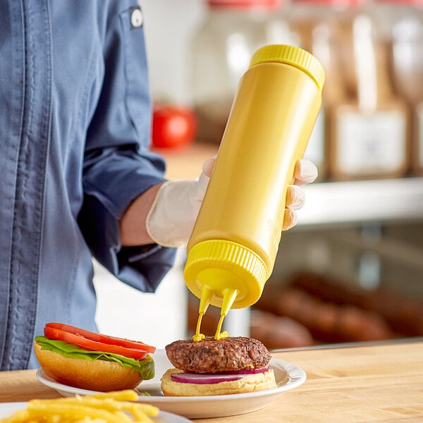 A hand using a Vollrath yellow squeeze bottle to put mustard on a burger.