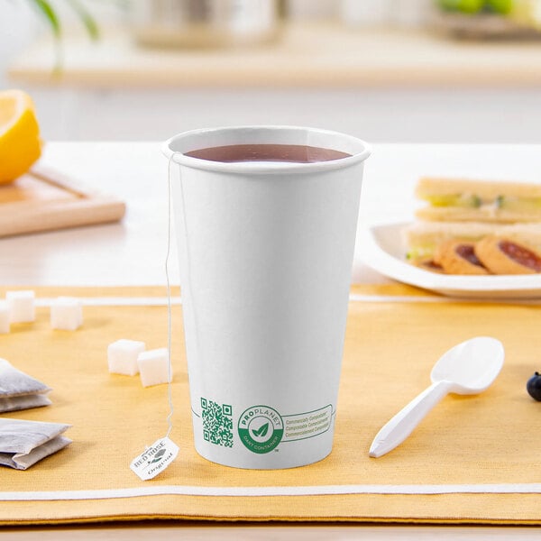 A white Solo ProPlanet compostable paper hot cup with a lid and a white spoon on a wood table with a cup of tea and a tea bag.