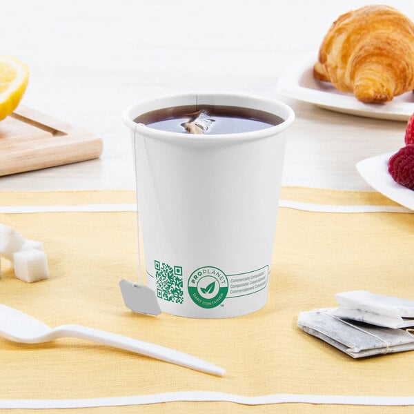 A Solo white compostable paper hot cup filled with tea on a table with a croissant and fruit.