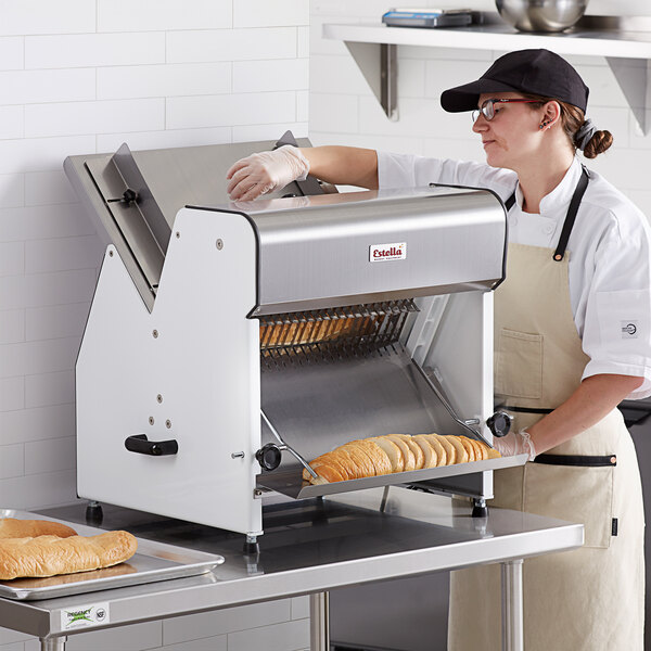 A person in a white coat and gloves using an Estella countertop bread slicer to slice bread.