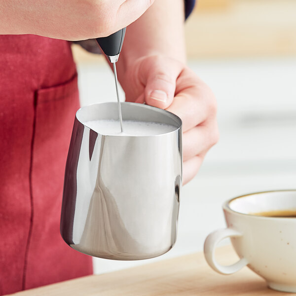 A person pouring white milk from a Tablecraft stainless steel frothing pitcher into a cup of coffee.