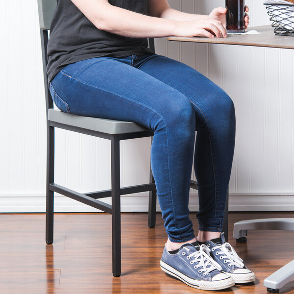 A woman sitting in a BFM Seating Memphis sand black steel side chair with gray wooden back and seat at a table.