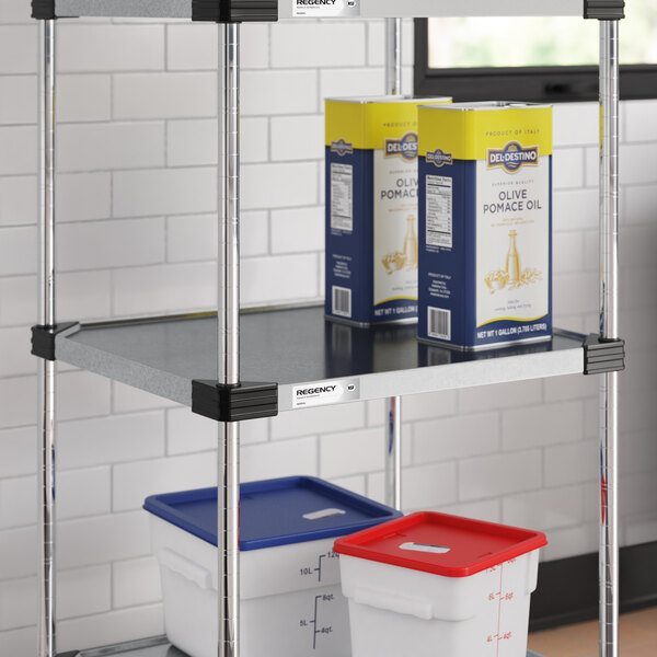 A metal shelving unit with two large containers on the lower shelf and two boxes of olive pomace oil on the middle shelf, set against a white tiled wall.