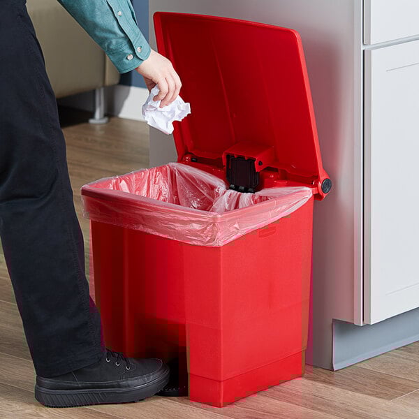 A person throwing a paper into a red Rubbermaid step-on trash can.