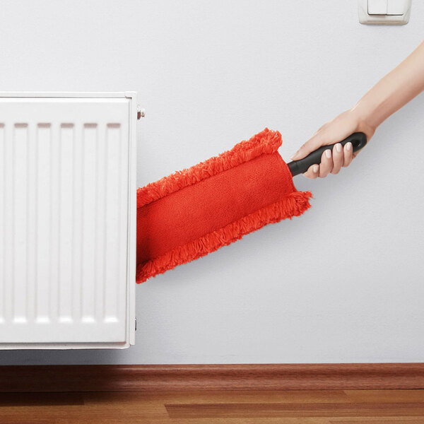 An OXO Good Grips microfiber under appliance duster with a red cleaning head and black handle being used to clean under a radiator.