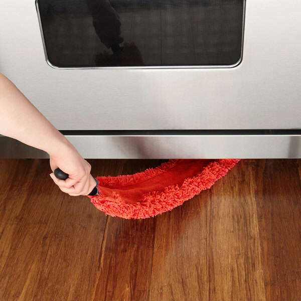 An OXO Good Grips 31 1/4-inch microfiber under appliance duster being used to clean beneath a stainless steel appliance on a wooden floor.