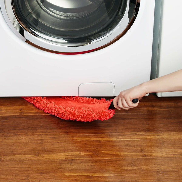 A red microfiber under appliance duster being used to clean beneath a washing machine.