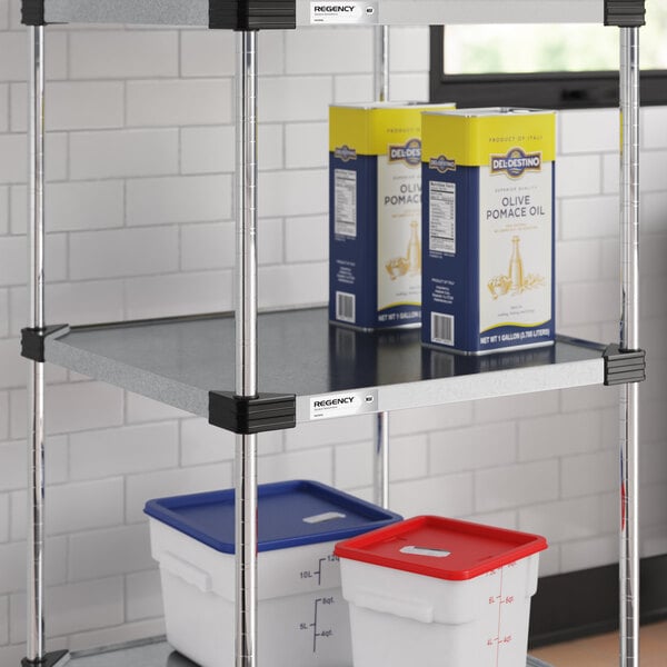 A metal shelving unit with two large boxes of olive pomace oil on the middle shelf and two plastic storage containers with colored lids on the lower shelf.
