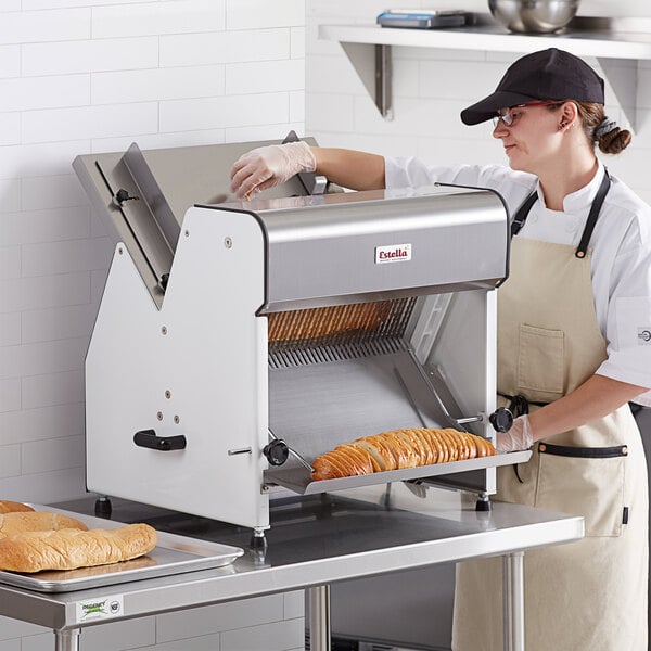 A woman in a white and black apron and hat slicing bread with an Estella countertop bread slicer.