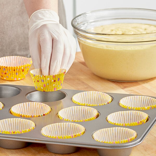 A person wearing gloves placing a yellow Wilton cupcake wrapper with colorful polka dots into a muffin tin.