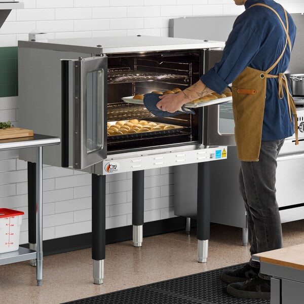 A single deck, full size electric convection oven with a stainless steel exterior, shown in use by a person placing a tray of baked goods inside.