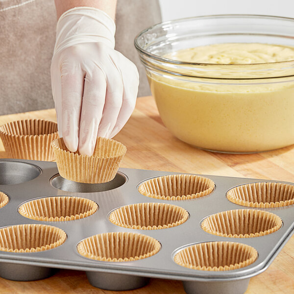 A gloved hand putting a Wilton Kraft unbleached cupcake wrapper into a muffin tin.