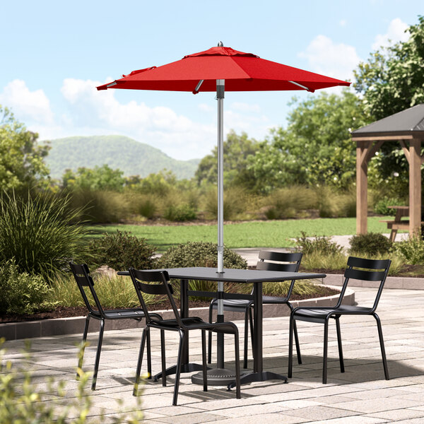 A red Lancaster Table & Seating umbrella on a table with chairs on a stone patio.