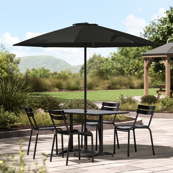 A black Lancaster Table & Seating umbrella over a table with chairs on a stone patio.